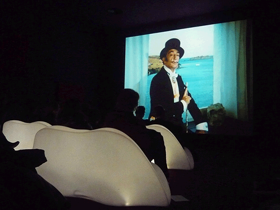 animation of a cinema audience watching Salvador Dali wearing a mourning suit and a top hat, raising his hand and gesticulating on a continous loop
