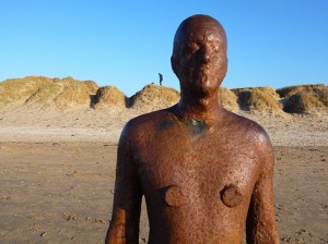 cast-iron, life-size figure against blue sky with sand dunes in the background.