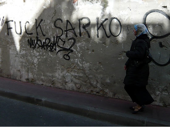 woman in an arabic head dress walkes past a wall on which is writtenl with "fuck sarko" written in large letters
