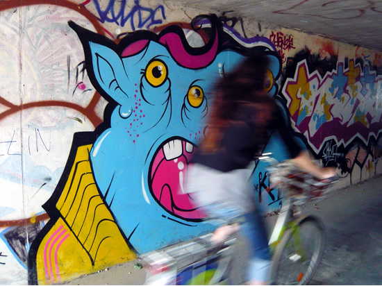 girl cycles past graffiti of a blue alien with three eyes