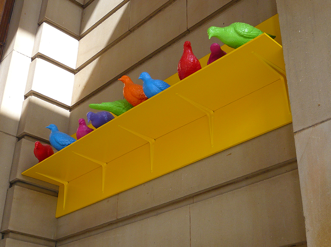sculpture of brightly coloured pigeons on a bright yellow, metal shelf against a sandstone block wall