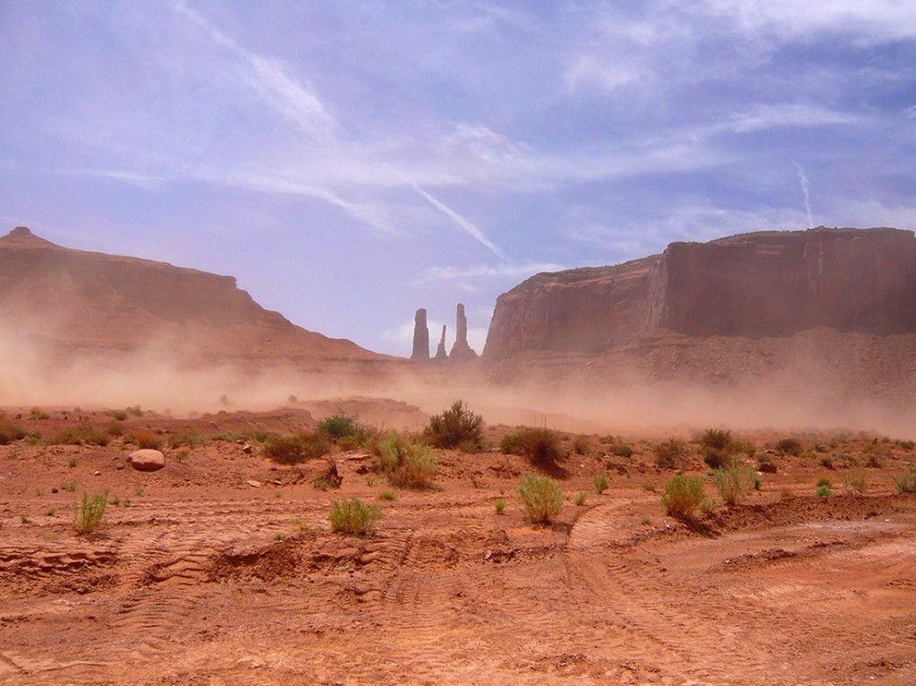 dust storm aproaching across the desert scrub land, tall cliffs and and three rock spires in the distance of monument valley
