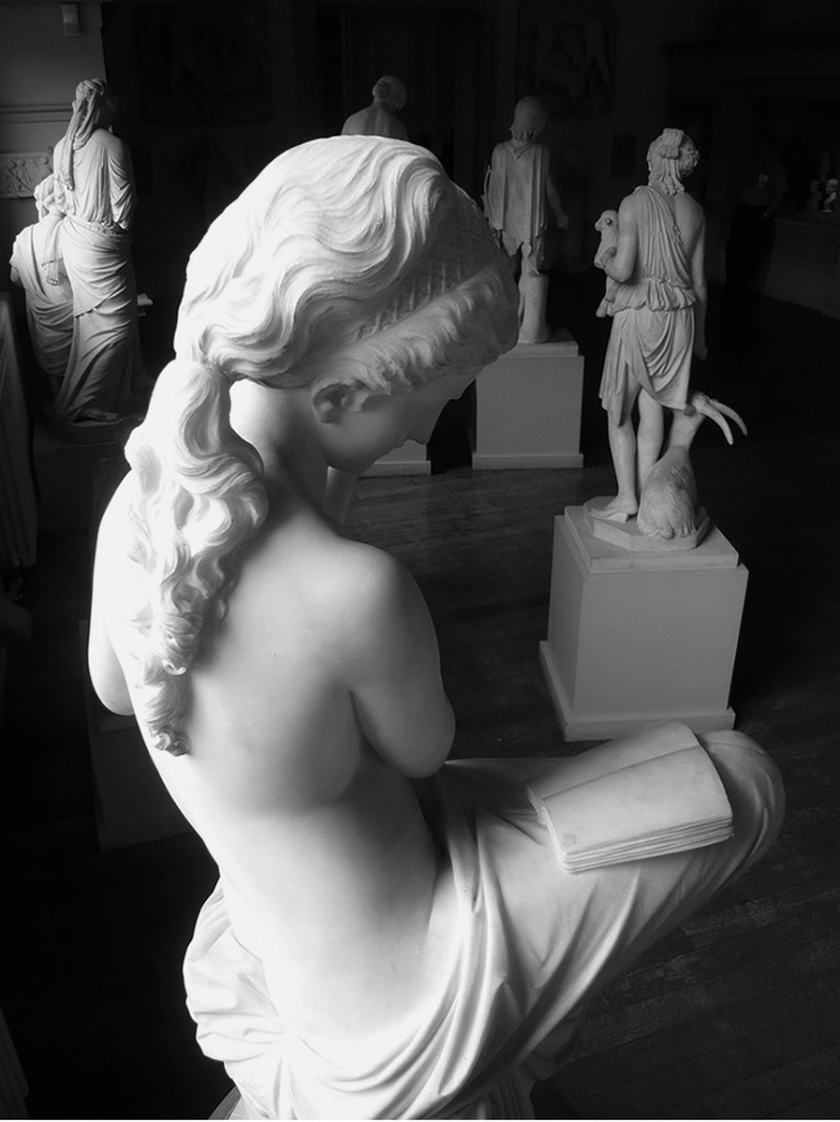 black and white photo of marble sculptures on plinths. in the fore ground a female figure reading