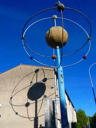 rusting sculpture of an "orrery" or planetary model (representing spheres in orbit) casting a shadow on the side of a house