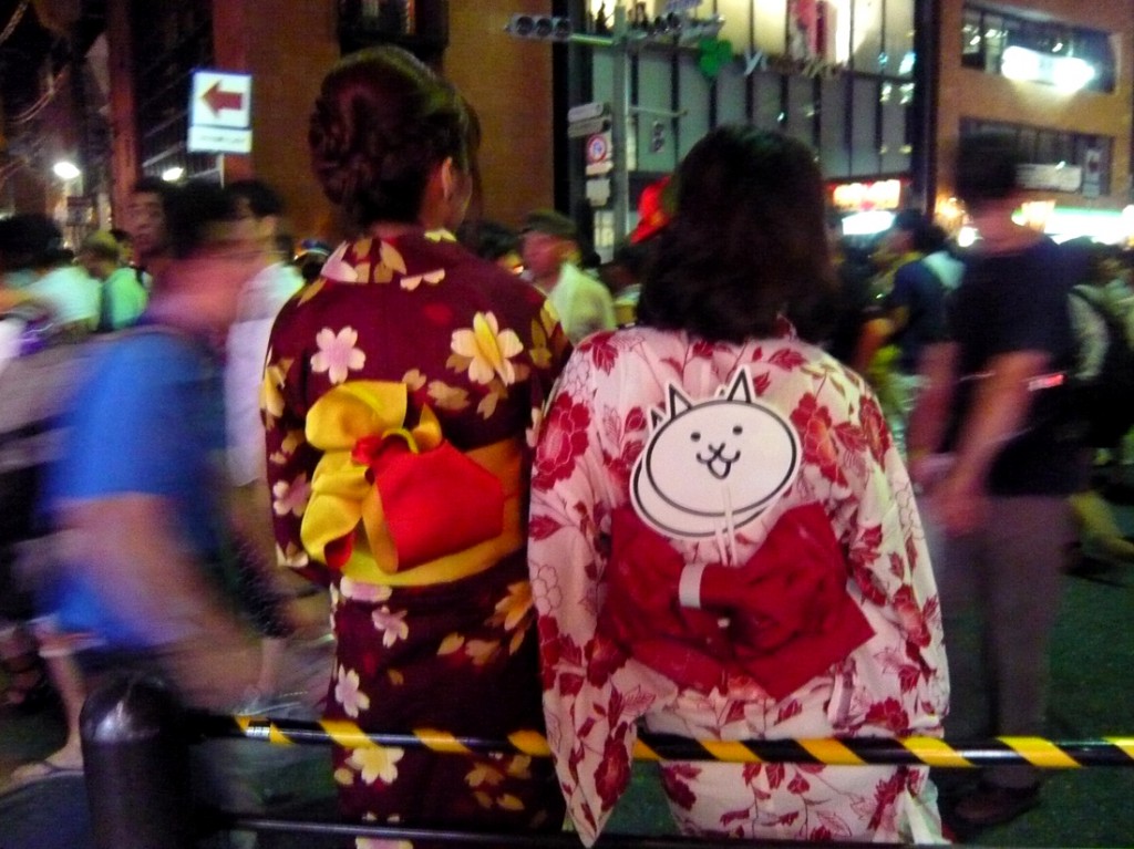 two young women wearing brightly coloured yukata (kimono) resting against a railing backs to camera as they watch festival goers in a blured street scene at nightured night