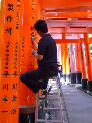 man painting black Japaneese characters on a red vermillion arh