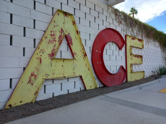 Large metal letters peeling yellow and red paint on a white brick wall spelling-out the word ACE
