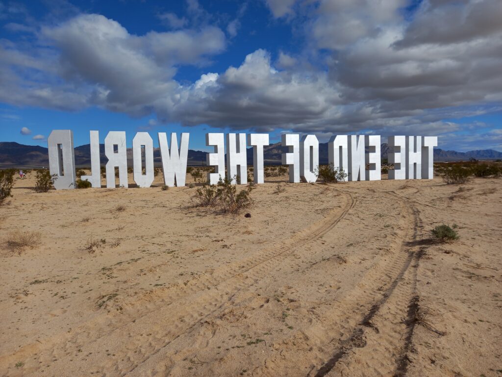 Large upright silver Hollywood-sign-style letters spelling out THE END OF THE WORLD. View from behind the letters showing the words backwards. Letters placed in a desert landscape. 