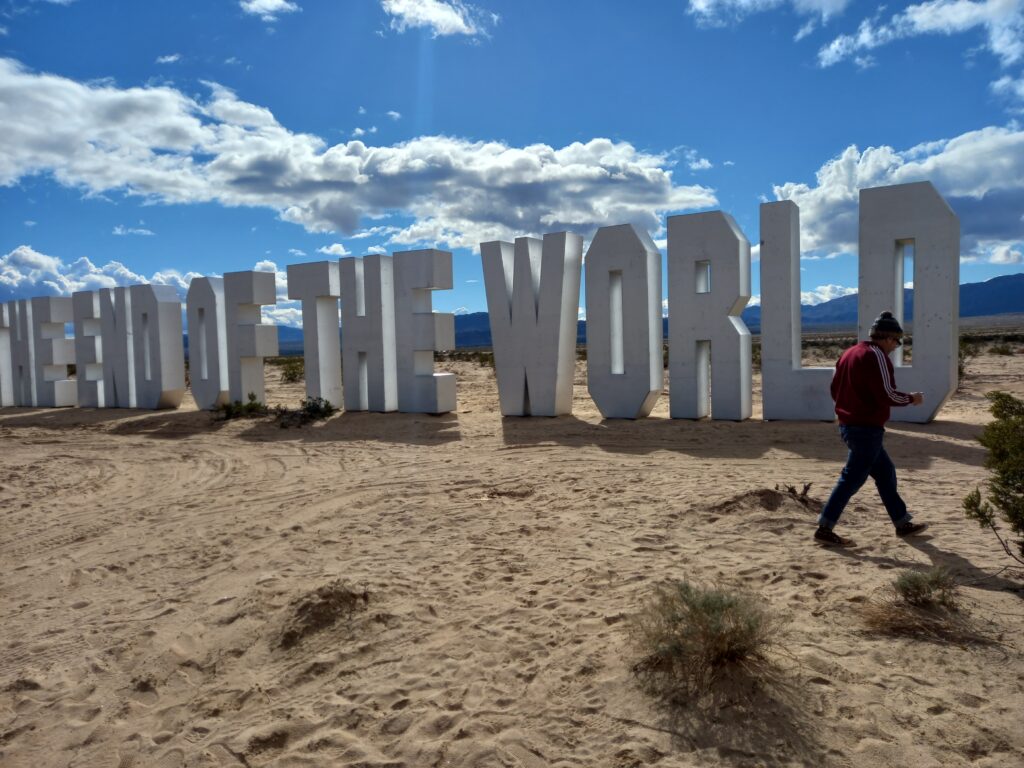 Man walks in-front of Large upright silver Hollywood-sign-style letters spelling out THE END OF THE WORLD.