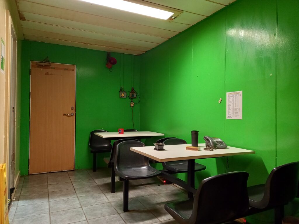 Plastic tables and chairs in and empty room. Walls painted bright green. Overflowing ashtrays and plastic gray telephone on table fixed to the floor. 